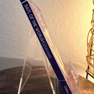 May include: A hardcover book titled "Bats of the World" by Graham, displayed on a clear acrylic book stand. The book has a dark blue spine and the Golden Books logo. The book is angled, showcasing the title and author.