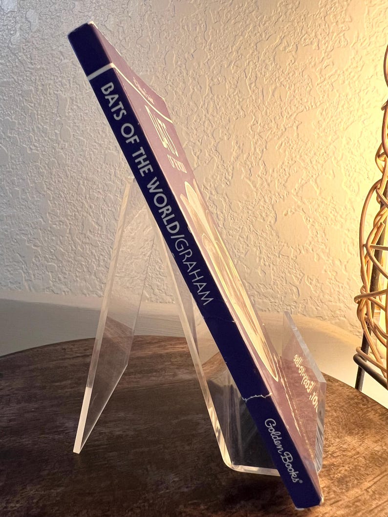 May include: A hardcover book titled "Bats of the World" by Graham, displayed on a clear acrylic book stand. The book has a dark blue spine and the Golden Books logo. The book is angled, showcasing the title and author.