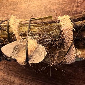 May include: A bundled sage smudge stick with a clear quartz crystal attached. The sage is wrapped with twine, black wire, and a woven tan fabric. The bundle is on a wooden surface.