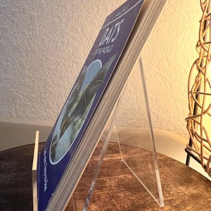 May include: A book titled "Bats of the World" is displayed on a clear acrylic stand. The book has a blue cover with white text and a bat illustration. The stand is angled, and the book is open, revealing the pages.