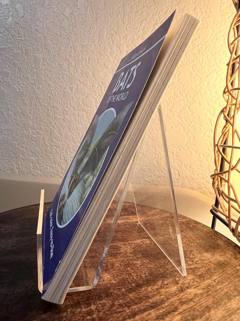 May include: A book titled "Bats of the World" is displayed on a clear acrylic stand. The book has a blue cover with white text and a bat illustration. The stand is angled, and the book is open, revealing the pages.