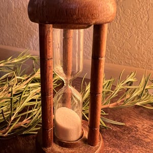 May include: A vintage hourglass with a wooden frame and a clear glass chamber. The hourglass is filled with light-colored sand. The wooden frame has a warm, brown tone. The hourglass is a decorative item.