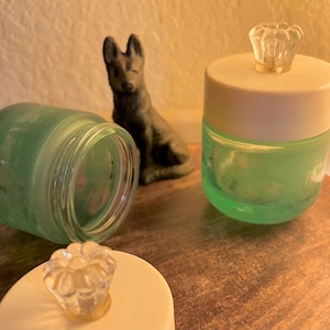 May include: Two small, round, light green glass jars with white lids. Each lid features a clear, flower-shaped glass knob handle. A small, dark gray, seated dog figurine is positioned between the two jars, one of which is lying on its side.