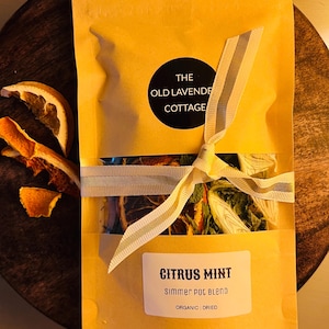 May include: A yellow paper bag tied with a striped ribbon, containing a Citrus Mint simmer pot blend. The bag has a black circle with "THE OLD LAVENDER COTTAGE" printed on it. Dried orange slices are next to the bag. The label says "ORGANIC | DRIED".