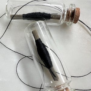 May include: Two clear glass bottles with cork stoppers. Each bottle contains a bundle of black thread wrapped around a wooden dowel. Black thread extends from the bottles. The bottles are displayed on a white surface.