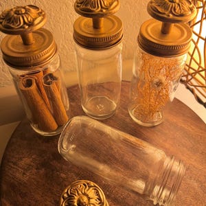 May include: Four clear glass jars with gold-toned metal lids featuring a raised floral knob design. One jar contains brown cinnamon sticks, one contains dried yellow foliage, and two are empty. The lids are screw-top style with a metallic finish.