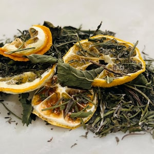 May include: Close-up of a pile of dried herbs and citrus slices. The image features dried lemon slices, green herbs, and rosemary. The ingredients are arranged on a white surface, suggesting a culinary or herbal preparation.