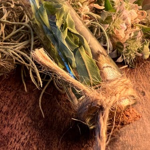 May include: A clear glass vial filled with green leaves, sealed with a cork and tied with twine. The vial rests on a brown surface, with dried flowers and greenery in the background. The overall aesthetic is natural and rustic.