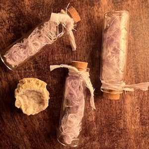 May include: Three clear glass vials with cork stoppers, each containing tangled light purple thread and tied with a white fabric ribbon, accompanied by a single rough, circular geode stone with a crystalline interior and a tan exterior.