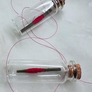 May include: Two small glass bottles with cork stoppers. Inside each bottle is a small wooden stick wrapped with bright red thread. Red thread extends from the bottles, creating loops.