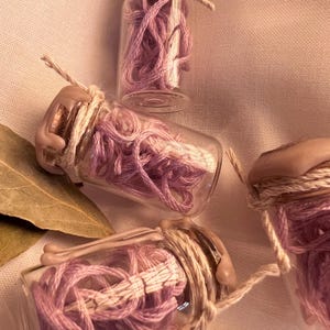 May include: Small glass bottles filled with pink thread, sealed with wax and twine. The bottles are arranged on a light pink fabric, with a dried leaf visible in the background. The bottles are a decorative item.