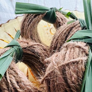 May include: Close-up of several brown rope wreaths, each adorned with green leaves tied around the wreath. The wreaths are arranged in a pile, with some visible in the background. The natural materials suggest a rustic, eco-friendly theme.