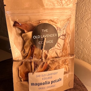 May include: A kraft paper bag with a clear window containing dried magnolia petals. The bag is sealed with a twine tie and features a round label with "The Old Lavender Cottage" and a rectangular label with "magnolia petals" and "dried | organic".