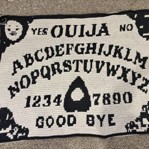 May include: A black and white Ouija board blanket. The blanket features the alphabet, numbers, and the words "YES", "NO", and "GOOD BYE". The design includes a planchette shape and decorative elements.