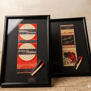 May include: Two framed vintage matchbook covers in black frames. One has red and white concentric circles with text, the other a red and black illustration with text. Several matches with red tips are placed in front of the frames.