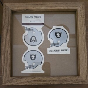 May include: Framed vintage Oakland Raiders and Los Angeles Raiders matchbooks. The matchbooks feature helmet designs with the Raiders logo. The frame is made of light brown wood. The text "OAKLAND RAIDERS" and "LOS ANGELES RAIDERS" are visible.
