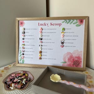 Könnte beinhalten: Ein gerahmtes Schild mit dem Text "Lucky Scoop" und einer Liste von Artikeln mit Illustrationen. Eine kleine Schale mit bunten Charms und ein graues Tablett mit einem dekorativen Löffel befinden sich im Vordergrund. Das Schild hat florale Akzente.