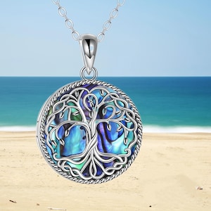 May include: A silver-toned necklace with a round pendant featuring a tree of life design. The pendant has a blue, green, and purple iridescent abalone shell background. The necklace is displayed against a beach and ocean backdrop.