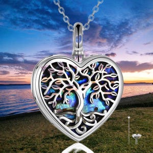 May include: A silver heart-shaped pendant necklace with a tree of life design. The pendant features a mother-of-pearl inlay with blue and green hues. The necklace hangs against a scenic background of a sunset over the water.