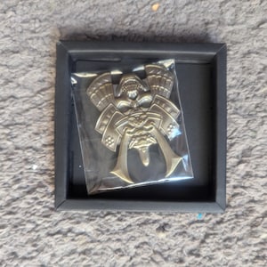 Puede incluir: Un emblema metálico de casco de samurái, presentado en un marco cuadrado negro. El emblema está dentro de una funda de plástico transparente. El casco de samurái tiene detalles intrincados, incluyendo una máscara facial y elementos decorativos. El marco está sobre una superficie texturizada de color claro.