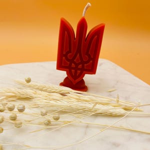 Puede incluir: Una vela roja con forma de tridente, colocada sobre una superficie de mármol blanco. La vela tiene una mecha y está rodeada de espigas de trigo secas y pequeños elementos decorativos beige redondos. El fondo es de color naranja liso.