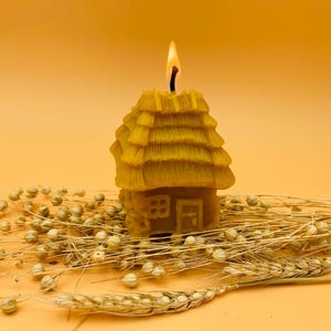 Puede incluir: Una vela encendida de cera de abeja amarilla con forma de casita con tejado de paja. La vela está sobre trigo seco y pequeñas vainas redondas. El fondo es de color naranja cálido.