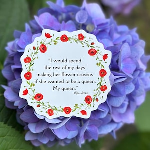 Puede incluir: Una tarjeta blanca, con bordes festoneados, con un borde floral de amapolas rojas y hojas verdes, sobre un fondo de flores de hortensia moradas. La tarjeta presenta la cita: "Pasaría el resto de mis días haciéndole coronas de flores si ella quisiera ser reina. Mi reina." -Kai Azer.