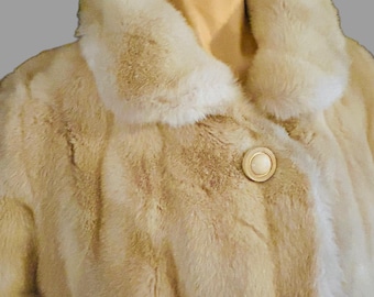 May include: A beige fur coat with a large collar and a single button closure. The button is a large, round, cream-colored button with a gold rim.