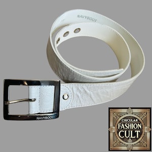 May include: White leather belt with a silver buckle. The buckle has the text "NAVYBOOT" engraved on it. The belt is folded in half and the text "NAVYBOOT" is visible on the top of the belt.