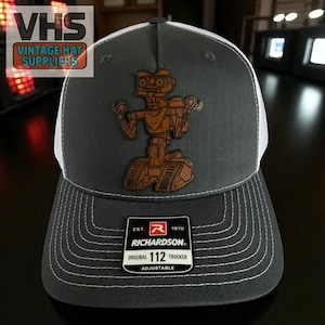 May include: A gray trucker hat with a white mesh back and a brown leather patch featuring a robot design. The hat has a curved brim with white stitching and a Richardson patch. The text "VHS Vintage Hat Suppliers" is in the upper left corner.