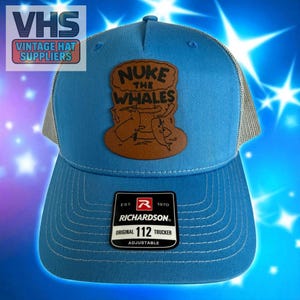 May include: A blue trucker hat with a mesh back and a curved brim. The hat features a brown leather patch with the words "NUKE THE WHALES" and a cartoon graphic. The hat has a Richardson 112 Trucker tag.