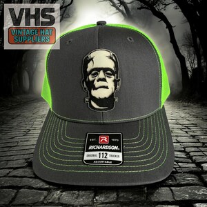 May include: A gray and neon green Richardson 112 trucker hat featuring a Frankenstein's monster patch. The hat has a curved brim and adjustable closure. The hat has the text "VHS Vintage Hat Suppliers".