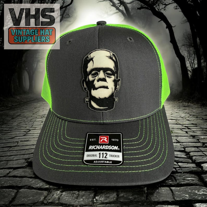 May include: A gray and neon green Richardson 112 trucker hat featuring a Frankenstein's monster patch. The hat has a curved brim and adjustable closure. The hat has the text "VHS Vintage Hat Suppliers".