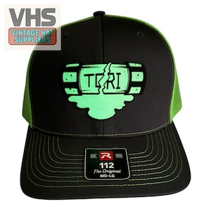 May include: A black baseball cap with bright lime green mesh back panels and lime green stitching on the brim. The front features a graphic of a cracked canister with the text "TCRI" in black, with a lime green liquid spill design underneath.