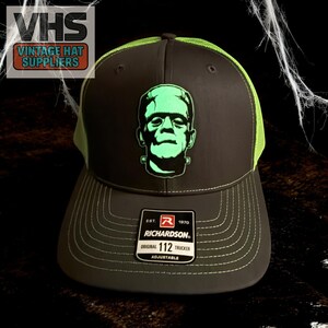 May include: A dark gray trucker hat with a neon green mesh back and brim. The front of the hat features a green graphic of Frankenstein's monster. The hat has a Richardson 112 tag and the text "VINTAGE HAT SUPPLIERS".