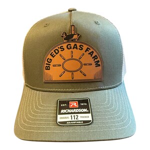 May include: An olive green trucker hat with a mesh back and a tan leather patch on the front. The patch features a graphic of a duck and the text BIG ED'S GAS FARM. A black sticker on the brim reads RICHARDSON ORIGINAL 112 TRUCKER EST. 1970.