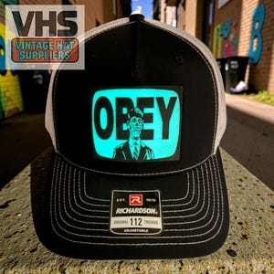 May include: Black and white trucker hat with a white mesh back and a curved brim. The front features a rectangular patch with the word "OBEY" in white letters and a graphic of a man. The hat is adjustable and has the Richardson 112 Trucker label.