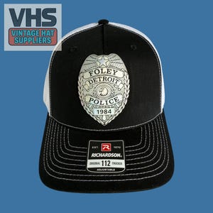 May include: Black and white trucker hat with a mesh back. The front features a police badge with the text "FOLEY DETROIT POLICE 1984". The hat also has the Richardson brand label and the text "ORIGINAL 112 TRUCKER".