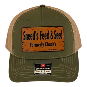 May include: An olive green trucker hat with a tan mesh back and a rectangular brown patch on the front. The patch features a wood grain texture and black text that reads Sneed's Feed & Seed Formerly Chuck's. A black Richardson 112 label is attached.
