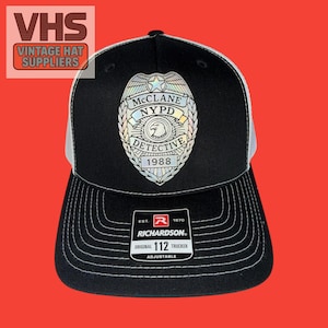 May include: Black and white trucker hat with a silver NYPD Detective badge. The hat features the text "McClane NYPD Detective 1988" and the Richardson brand label. The hat is adjustable.