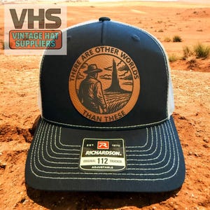 May include: A navy blue trucker hat with a white mesh back. The front features a leather patch with the text "THERE ARE OTHER WORLDS THAN THESE" around an illustration of a person and a tower. The hat has a curved brim and white stitching.