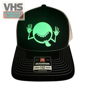 May include: A black and white trucker hat featuring a glow-in-the-dark graphic of a round, wide-eyed character with hands raised and tongue sticking out. The front panel displays the graphic, while a sticker on the brim reads Richardson 112.