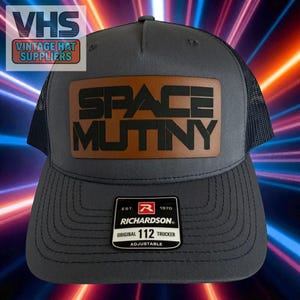 May include: A gray trucker hat with a mesh back and a brown leather patch. The patch features the words "SPACE MUTINY" in black block letters. The hat also has a "Richardson" tag and the text "VHS VINTAGE HAT SUPPLIERS".