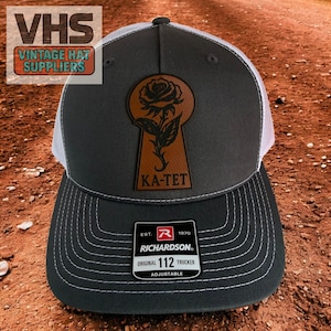 May include: A charcoal gray and white trucker hat with a leather patch featuring a rose in a keyhole design. The text "KA-TET" is below the keyhole. The hat has a curved brim and a Richardson 112 Trucker tag.
