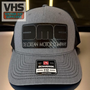 May include: Gray trucker hat with a black mesh back and a black DeLorean Motor Company logo. The hat has a curved brim and an adjustable strap. The Richardson tag reads "Original 112 Trucker".