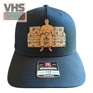 May include: A black trucker hat featuring a graphic of a person sitting on a floral patterned couch. The hat has a structured crown and a curved brim. A black and white sticker on the brim reads "EST. 1970 RICHARDSON ORIGINAL 112 TRUCKER ADJUSTABLE".