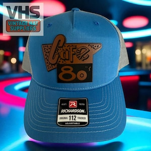 May include: A blue trucker hat with a mesh back and a brown leather patch that reads "Cafe 80's". The hat has a curved brim and an adjustable strap. The Richardson tag is visible.