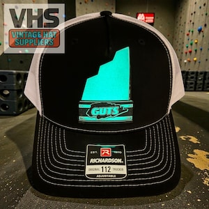 May include: Black and white trucker hat with a neon green graphic and the word "GUTS". The hat has a white mesh back and the text "RICHARDSON. ORIGINAL 112 TRUCKER ADJUSTABLE". The hat is from Vintage Hat Suppliers.