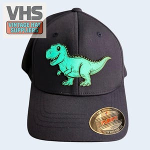 May include: Navy blue Flexfit youth hat featuring a smiling, light green dinosaur graphic. The hat has a curved brim and a gold Flexfit label. The text "VHS Vintage Hat Suppliers" is in the upper left corner.