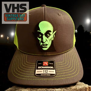 May include: A gray and neon green trucker hat with a green monster face patch. The hat has a curved brim and a mesh back. The text "VHS Vintage Hat Suppliers" is in the upper left corner. The hat is adjustable and has the text "Richardson Original 112 Trucker".